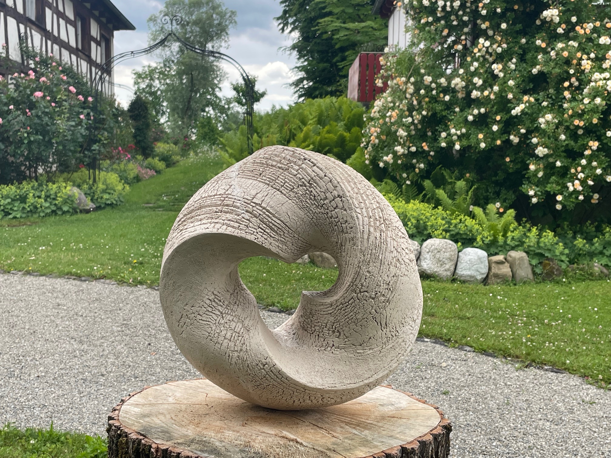 Skulptur „Alles im Fluss II“ von Sabine Classen – organische, kreisförmige Keramikform mit strukturierter Oberfläche, ausgestellt auf einem Baumstamm-Sockel im Garten des Schwandorfer Hofs in Salem am Bodensee, Keramikskulptur Verkaufsobjekt